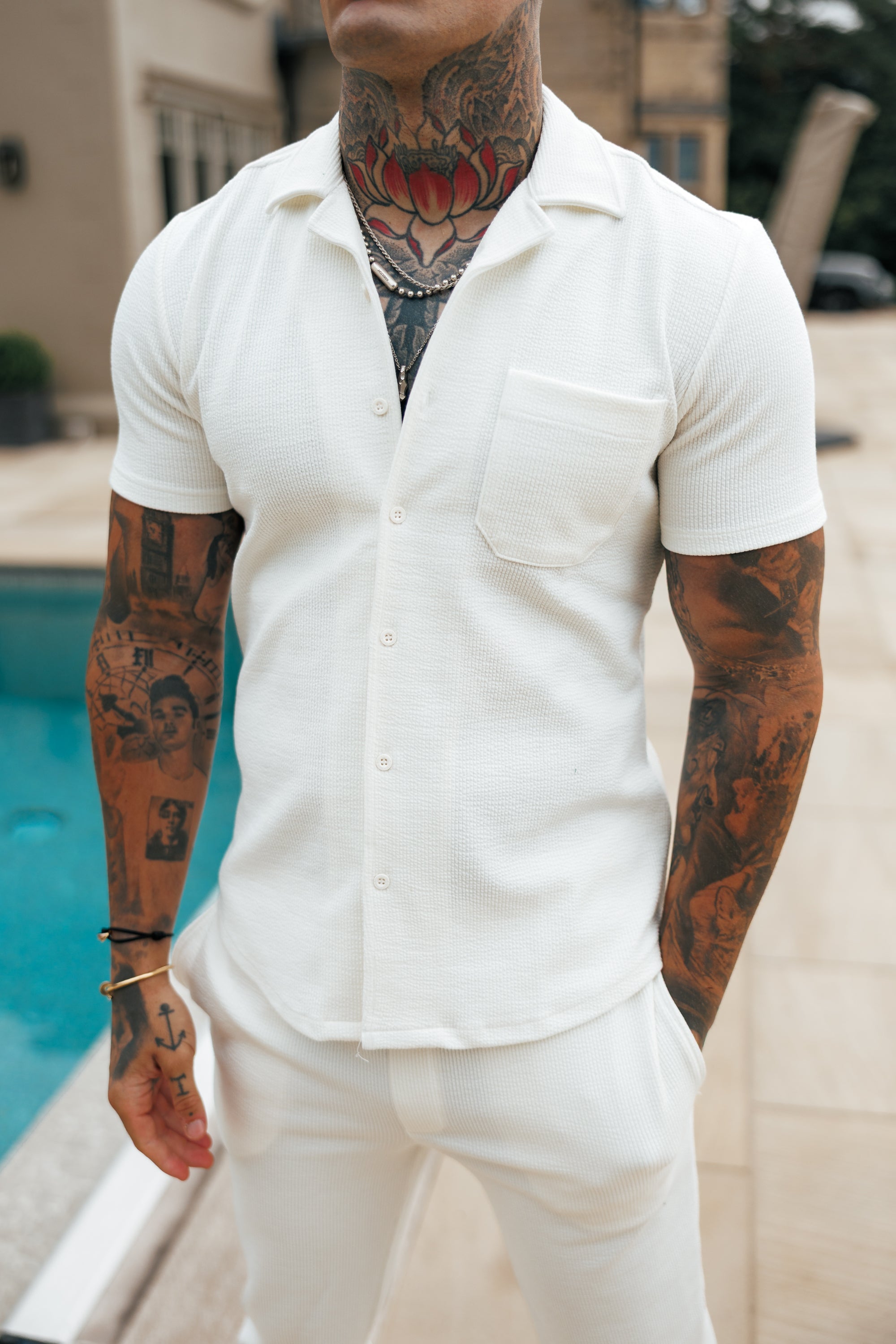 Man wearing a white short-sleeve shirt and pants with tattoos on his arms, standing by a pool.