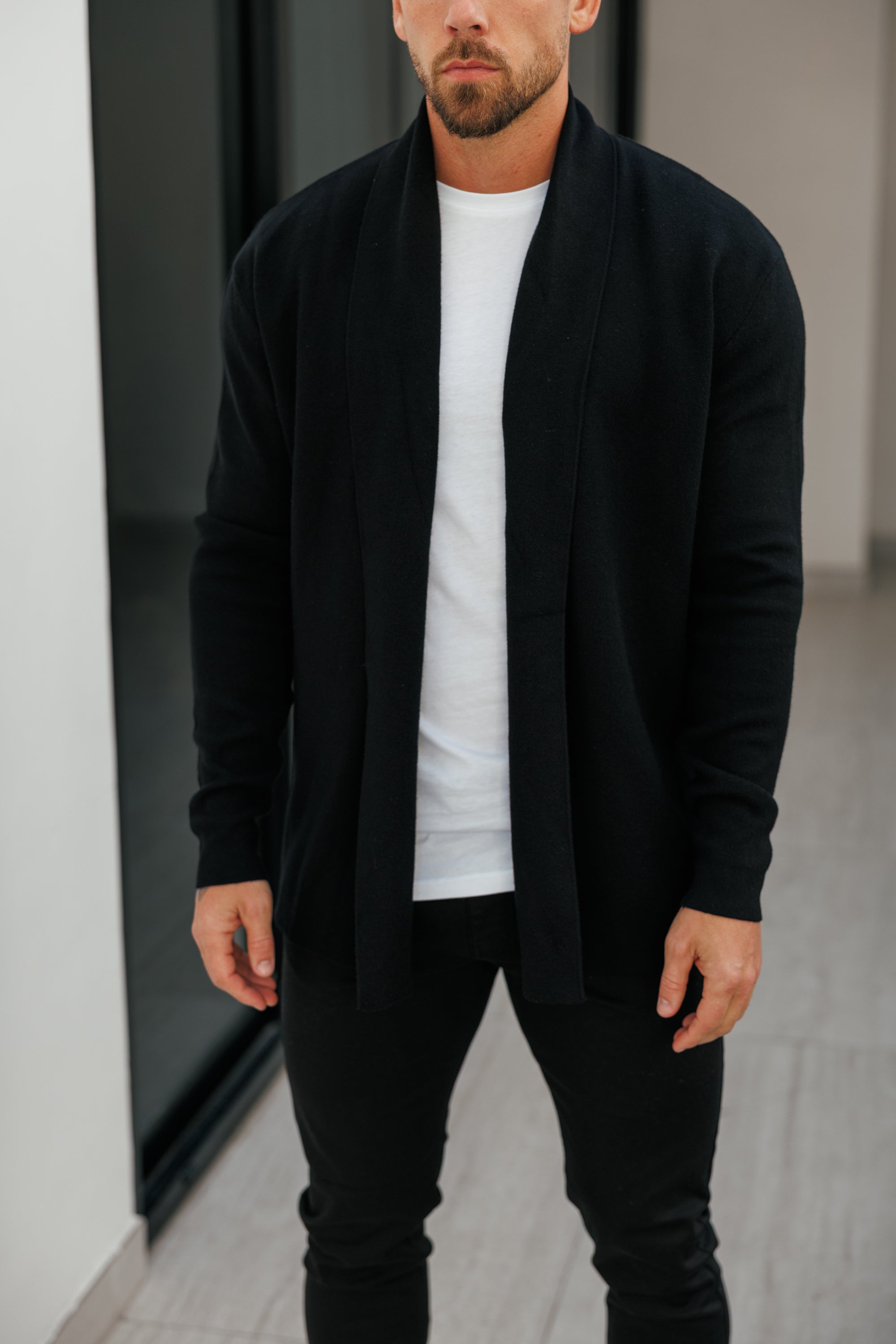 Man wearing a black cardigan over a white shirt and black pants in an indoor setting.