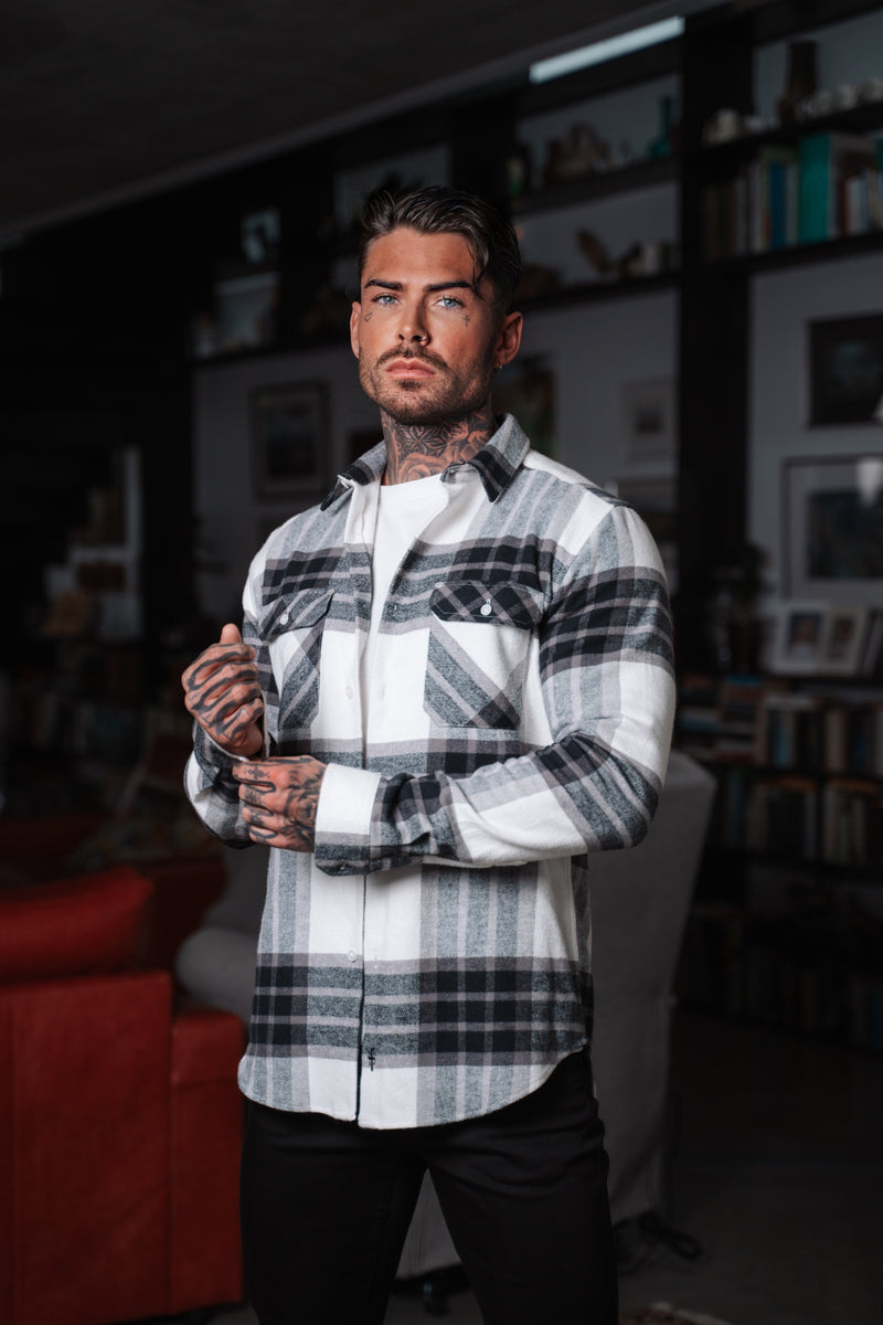 A man wearing an off white/grey checkered shirt with double pockets, standing in a room with bookshelves.