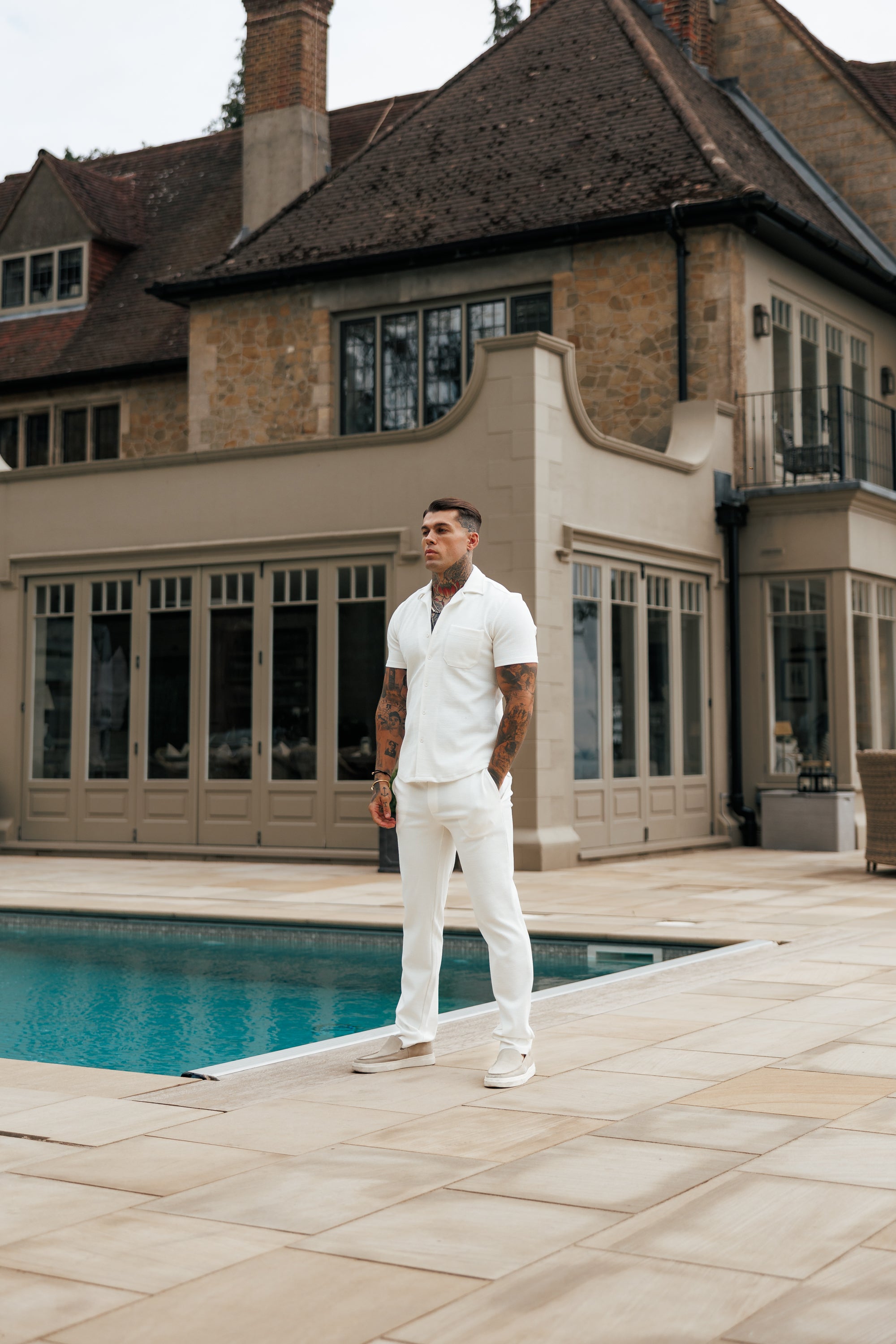 Man in a white outfit standing by a pool with a large house in the background