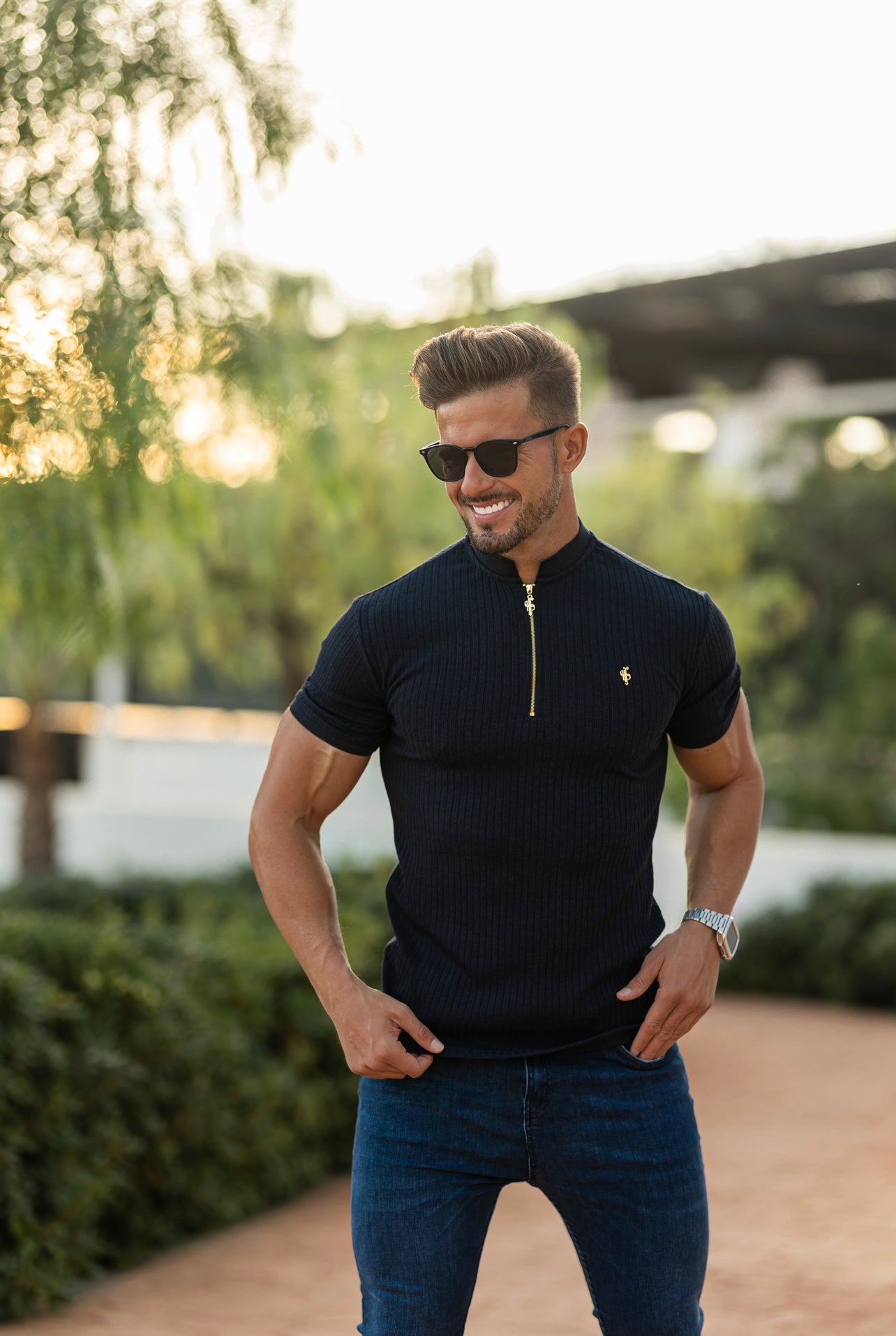A man wearing a navy ribbed zip grandad collar shirt with short sleeves, paired with sunglasses and standing outdoors.