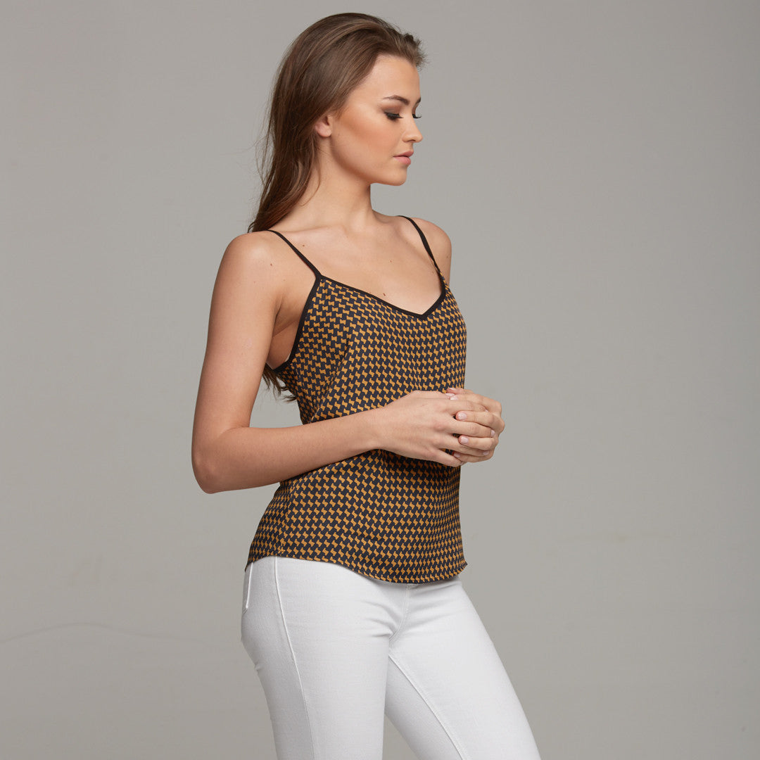 A female model wearing a brown and white printed camisole top with contrast black piping at the neckline and straps, paired with white trousers.